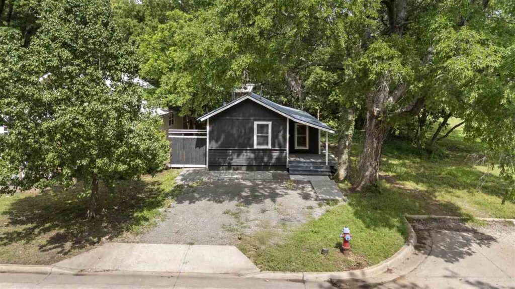 Charming black single-story home with front porch and gravel driveway surrounded by mature trees in Stillwater, Oklahoma