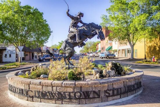 A large bronze statue of a cowboy riding a bucking bronco in a stone-walled roundabout in Historic Downtown Stillwater, OK, surrounded by green trees and local storefronts under a clear blue sky.
