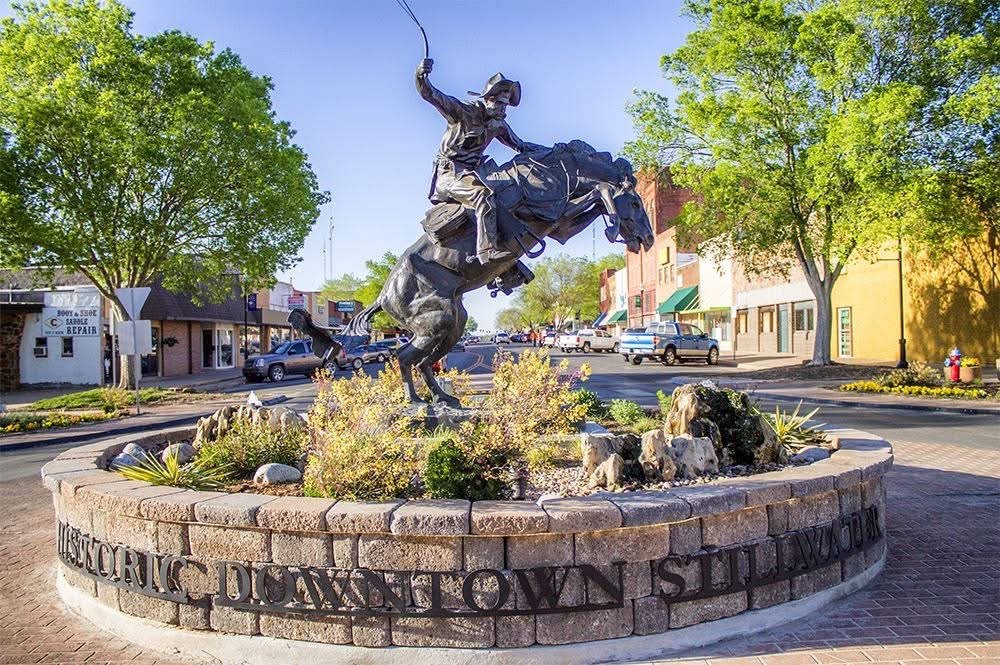A large bronze statue of a cowboy riding a bucking bronco in a stone-walled roundabout in Historic Downtown Stillwater, OK, surrounded by green trees and local storefronts under a clear blue sky.